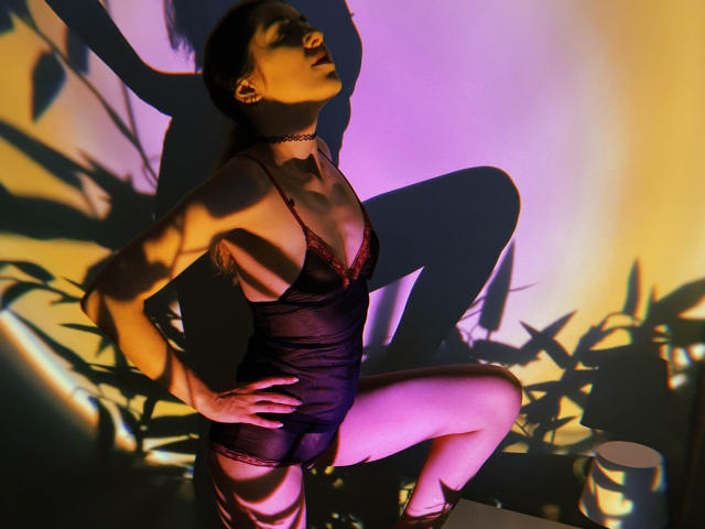 Decadance in black sheer lingerie with red lace, kneeling, hands on hips, choker, shadowed, purple and yellow light, legs apart, showing her ass, with a metal cup.