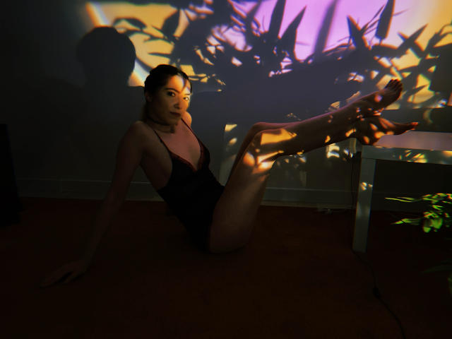 Decadance est assise par terre, vêtue d'un ensemble de lingerie en dentelle noire, les jambes relevées et ombragées par des feuilles de plantes, dans une pièce faiblement éclairée..