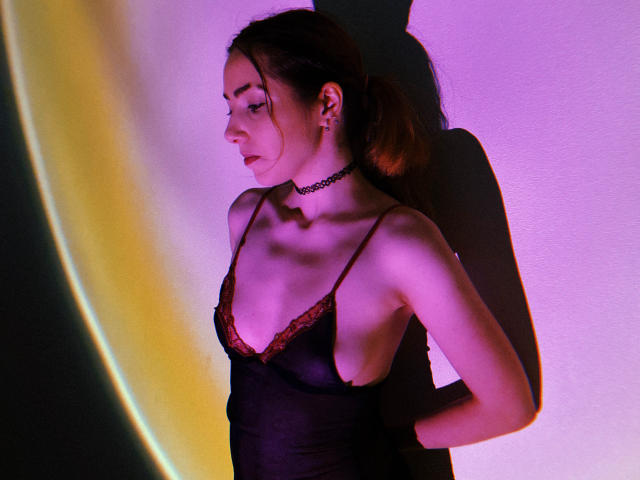 Decadance in black lingerie with red lace, choker, and pouty lips, arms behind back, shadow on wall, pink-purple light, showing small tits and slim, pale figure.