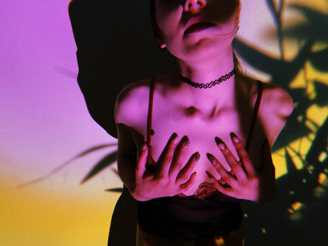 Decadance with her hands on her tits, wearing a black choker and lace bra, in purple and yellow lighting, shadow of her face behind her, painted nails.