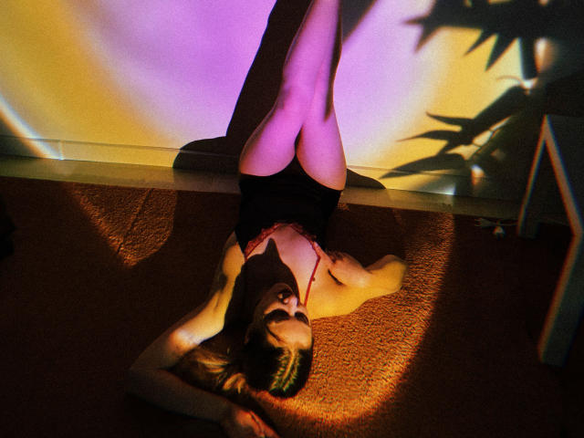 Decadance is lying on a carpet, legs up, wearing a red lace top, black bottoms, with purple and yellow shadows on her body and legs, arms resting above her head.