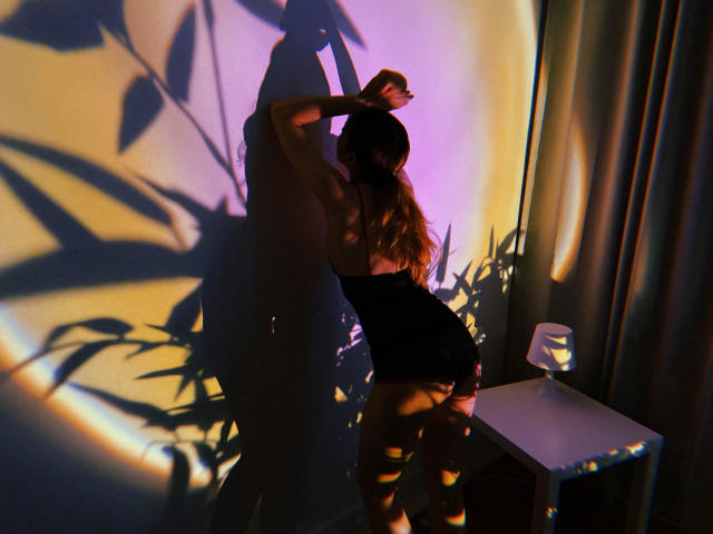Decadance with long brown hair, back to camera, wearing a black, tight, sleeveless top, arms raised, shadow of leaves on wall, standing next to a small table with a lamp.