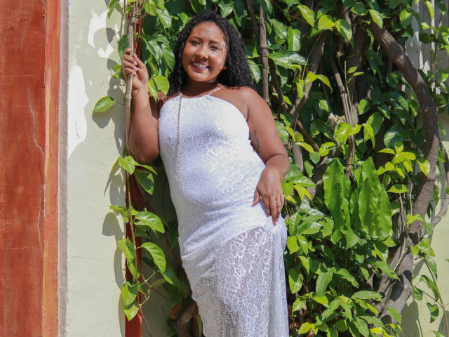 CaroldelaTorre, dark-skinned with curly black hair, wearing a white lace dress, standing against a green plant wall, smiling, right hand touching vine, left hand on hip.