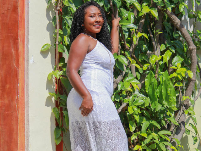 CaroldelaTorre, avec ses cheveux noirs bouclés, sa peau foncée, vêtue d'une robe blanche en dentelle, se tient près d'une plante verte et luxuriante, souriante, une main sur la plante, mettant en valeur ses fesses et ses seins..