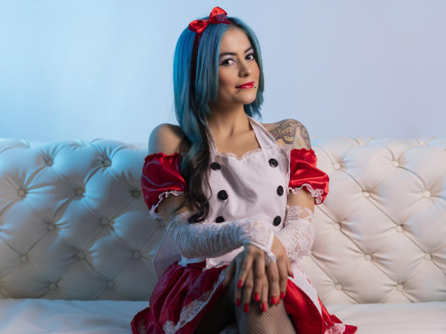 CandiceValo, avec des cheveux bleus, un nœud rouge, une tenue de femme de ménage rouge et blanche, des gants en dentelle blanche, assise sur un canapé blanc capitonné, montrant un tatouage sur son bras droit, souriante..