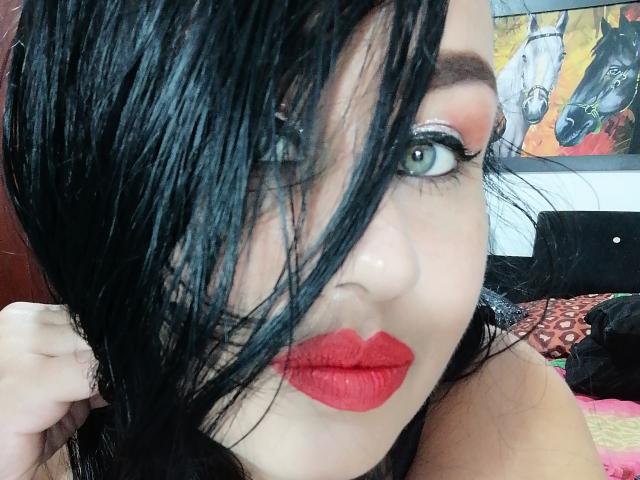 CathyCat has black, wet-looking hair covering one eye, bright red lipstick, and green eyes with bold eyeliner and pink eyeshadow, looking at the camera with a slight smile.