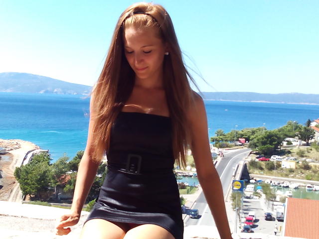DajlaSex hat langes, braunes Haar, kleine Brüste und trägt ein schwarzes, trägerloses Kleid mit einem Gürtel. Sie sitzt draußen, blickt nach unten, im Hintergrund ist ein blaues Meer und ein klarer Himmel zu sehen..
