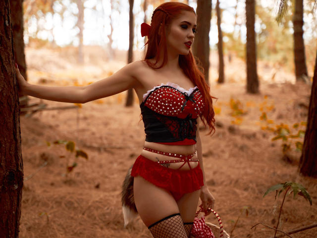 Maisy Warner, con i capelli rossi, indossa un top senza spalline a pois rossi e neri, biancheria intima in pizzo rossa e collant a rete, ed è in piedi in una foresta, tenendo un nastro rosso e bianco..