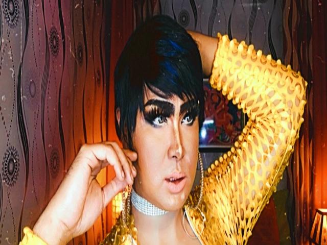 BornStarTS with short black hair, wearing a yellow crochet top, chandelier earrings, and a sparkly choker, raising one arm, looking seductively at the camera with dark, dramatic eye makeup.