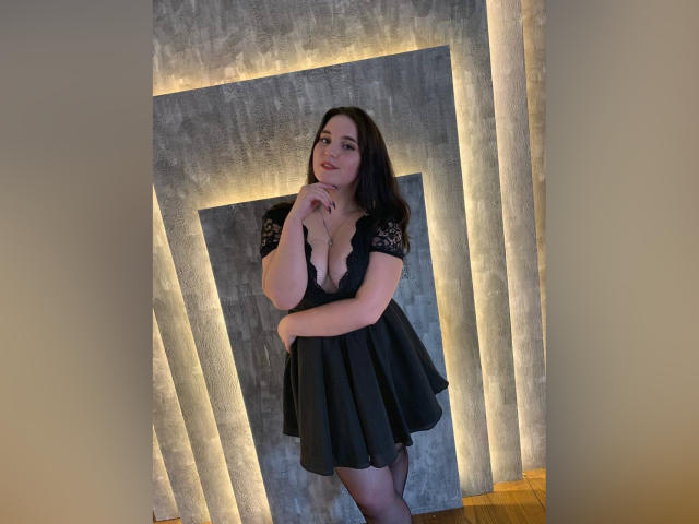 Lianna, avec ses cheveux foncés, portant une robe noire en dentelle avec un décolleté en V plongeant, debout, la main près de sa bouche, parquet en bois, mur en béton éclairé, séduisante, aux formes généreuses, sourire doux..