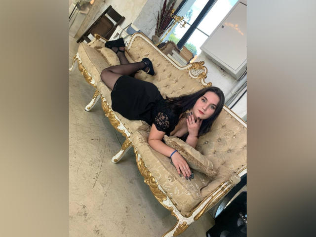 LiannaAh laying on a beige, gold-trimmed couch, wearing a black lace dress, black stockings, and black heels, with her tits slightly visible, and her ass raised.