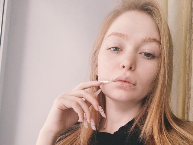 SweetAleksaX tiene la piel pálida, cabello largo y rojo, y lápiz labial rosa, con un dedo cerca de sus labios fruncidos, mostrando uñas manicuradas en tonos rosa y blanco, y parece ligeramente aburrida..