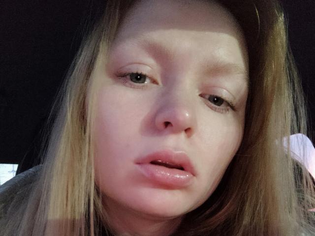 Het gezicht van SweetAleksaX is in beeld, met glanzende, licht open roze lippen, fonkelende ogen die wegkijken van de camera, en blond haar dat haar gezicht omlijst. Haar neus is klein en schattig..