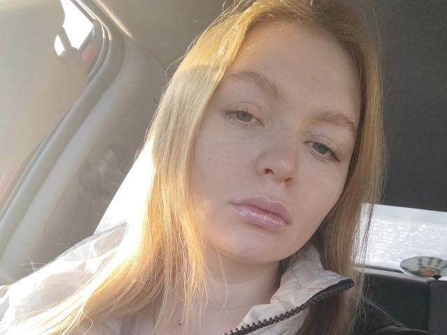 SweetAleksaX has pale skin, blonde hair, and shiny pink lips. She's wearing a white jacket, sitting in a car with sunlight on her face. Her expression is neutral.