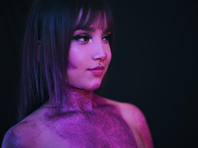 TamaraOlsen met paars glitter op haar gezicht, nek en borst, donker steil haar, een neusring en make-up, in een portret met paars en roze neonverlichting..