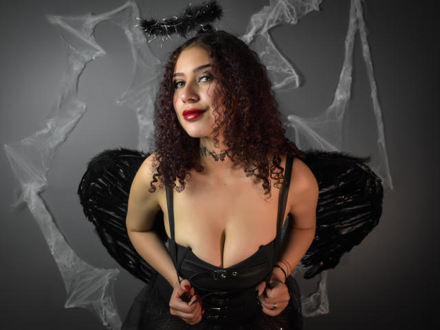 ValeryScot with curly brown hair, red lipstick, black dress, low cleavage, black wings, and black halo, holding dress, looking up, gray background. Boobs, wings, halo, dress.