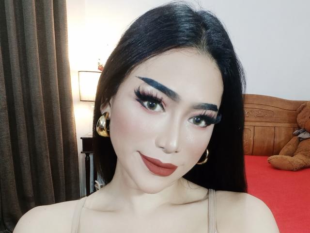 TransAsianSweetX has long black hair, thick black eyebrows, long eyelashes, red lipstick, and gold hoop earrings, wearing a beige top, with a slight smile.