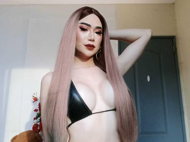 TransAsianSweetX mit langen, rosa Haaren, einem schwarzen, winzigen Bikini, der kleine Brüste zeigt, heller Haut, rotem Eyeliner, Reifenohrringen, einem angehobenen Arm, die in die Kamera schaut, eine Tür im Hintergrund..