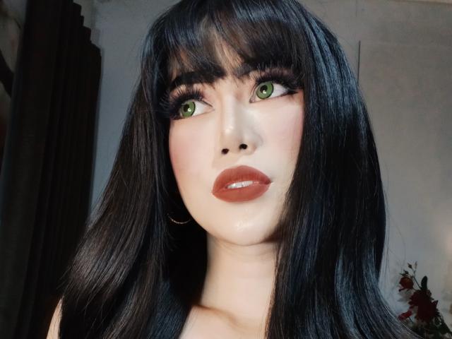 TransAsianSweetX tiene el cabello negro brillante, ojos verdes y labios rojos brillantes. Está mirando hacia un lado, con pestañas largas y gruesas y un rostro ligeramente sonrojado. No se ven los senos..