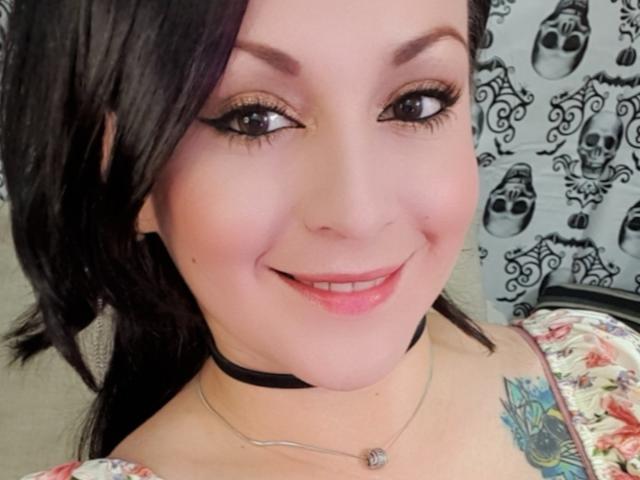 ElinaJhonson has dark hair, brown eyes with thick eyeliner, pink lipstick, black choker, floral dress, and a colorful tattoo on her right shoulder. She's smiling with pink lips.
