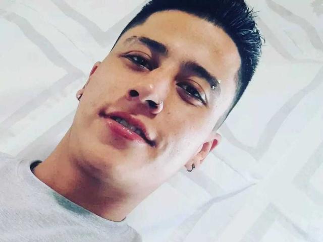 MarceLinio has short black hair, a nose ring, and a small earring. He's wearing a gray shirt, smiling slightly, with a small cut above his right eyebrow.