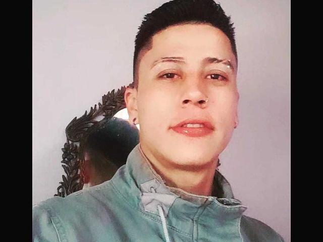 MarceLinio, con cabello negro corto, piel clara, una pequeña cicatriz sobre la ceja derecha, vistiendo una chaqueta verde, con la boca ligeramente abierta, un pendiente visible, mirando a la cámara..