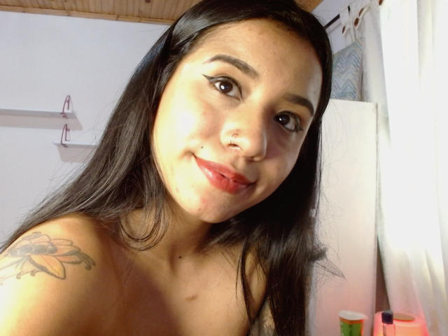 AshleyLover with long black hair, brown eyes, and red lipstick, smiling at the camera. She has a sunflower tattoo on her right shoulder and is topless.