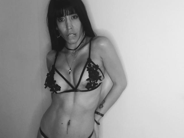 Black-and-white photo of SeleneBrigth with dark bangs, wearing a strappy, floral lace bralette, showing her small tits and flat stomach, standing with hands on her hips, looking at camera.