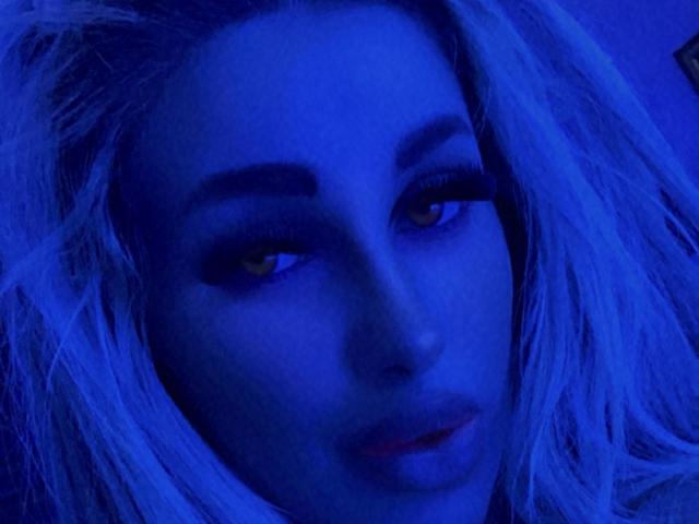 TanyaMonroe's face is lit in blue, showing yellow eyes with dark makeup, and blonde hair, looking slightly to the side with a subtle, sultry expression.