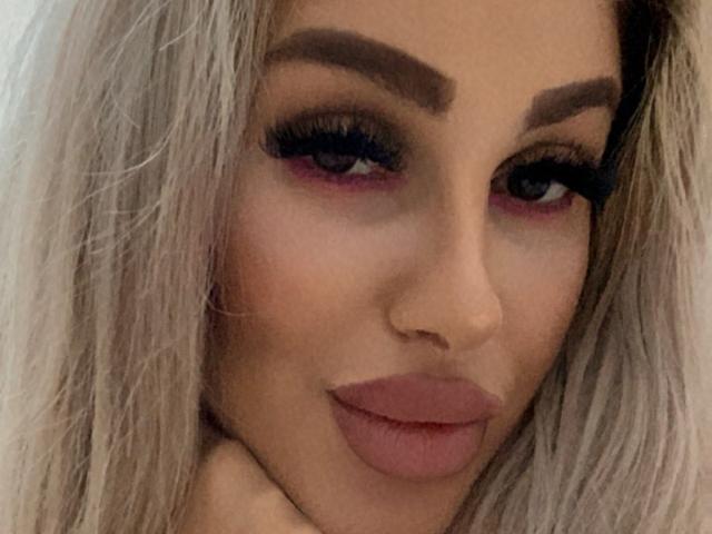 TanyaMonroe's face, showing her pouty ass-kissing lips, thick eyebrows, long blonde hair, dark eyeliner, and pink eyeshadow, with her hand under her chin, looking at the camera.