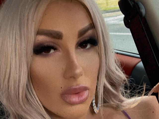 TanyaMonroe with platinum blonde hair, dark eyebrows, heavy eyeliner, long lashes, pink lipstick, and small moles on her face, sitting in a car.