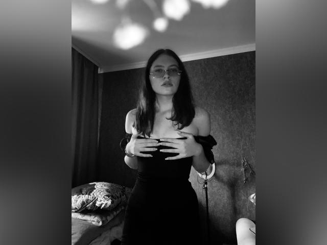 Bemahall con gli occhiali, capelli scuri e vestito nero con una spalla scoperta, mani che coprono il seno, in piedi in una stanza buia, con un'espressione seria, fotografia in bianco e nero, luce soffusa proveniente dall'alto..