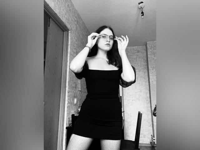 Bemahall, vêtue d'une robe noire et de bas résille, ajustant ses lunettes, debout dans un couloir, portant un bracelet, et affichant une attitude confiante. Noir et blanc, plan moyen, léger angle bas..