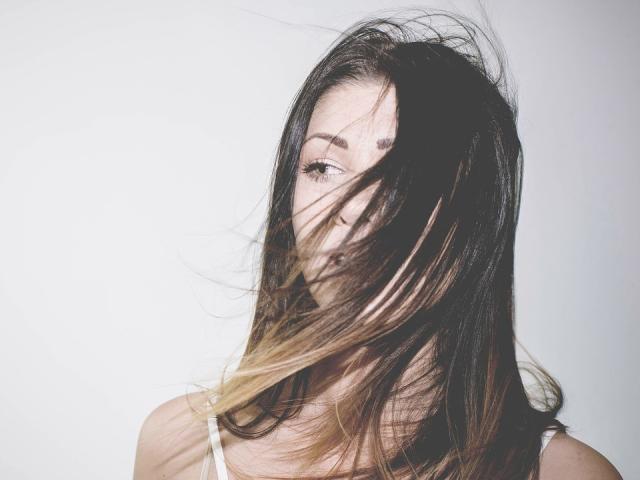 KaraWill's brown hair with blonde tips is blowing across her face, partially covering her eyes. She's wearing a white top, looking sexy with a subtle, confident smile.