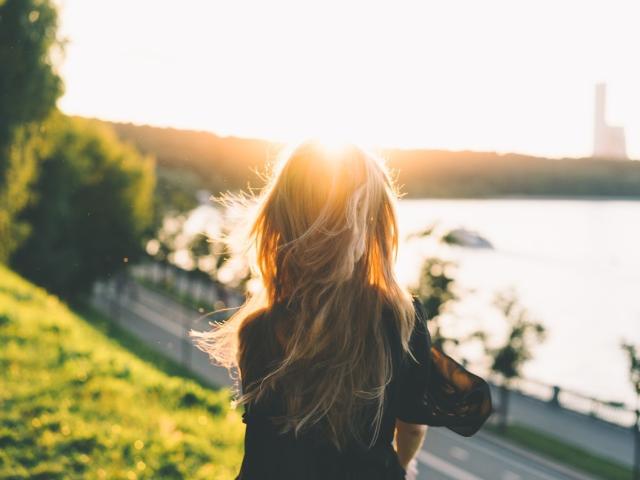 KaraWill está de vuelta, con su larga cabellera rubia, iluminada por el sol, vistiendo una blusa negra, de pie en una colina cubierta de hierba, mirando hacia un río, con una valla de metal y árboles al fondo..