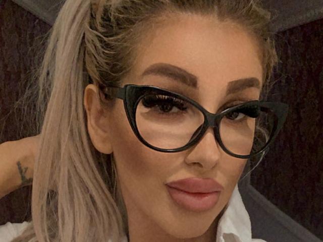 TanyaMonroe, con i suoi glutei prominenti, gli occhiali dalla montatura nera e la coda di cavallo bionda, ha un tatuaggio sul braccio sinistro e sta mostrando un'espressione compiaciuta e accigliata..