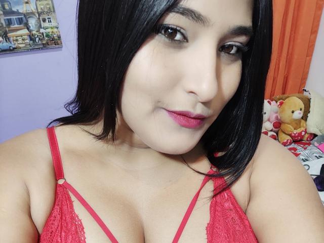 AndromedaAndSol, com cabelo castanho escuro, sutiã de renda vermelho, batom rosa e seios médios, sorrindo levemente, usando maquiagem discreta, em um selfie..