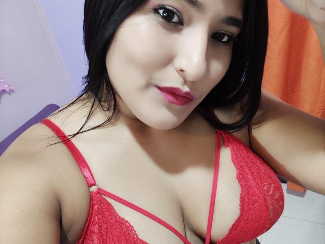 AndromedaAndSol tem pele clara, cabelo preto, um sutiã de renda vermelho com alças, que mostra seus seios, lábios rosados e olhos castanhos em uma selfie. Seu braço direito está levantado..