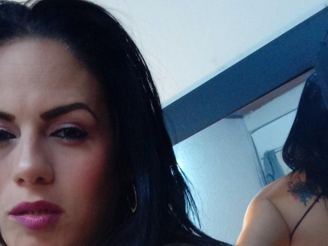 LizaRussell with black hair, pink eyeliner, and purple lipstick, showing her face and a reflection of her bare back with a small tattoo, in a mirror selfie.