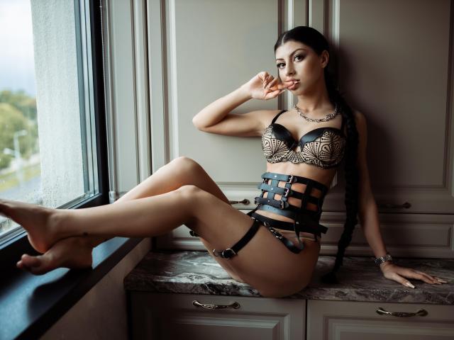 KateSparks, avec ses cheveux tressés foncés, portant un soutien-gorge en dentelle noire et une jarretière en cuir à brides, assise sur un rebord de fenêtre en marbre, mordant sa doigt, montrant ses seins et ses fesses, avec un regard séduisant..