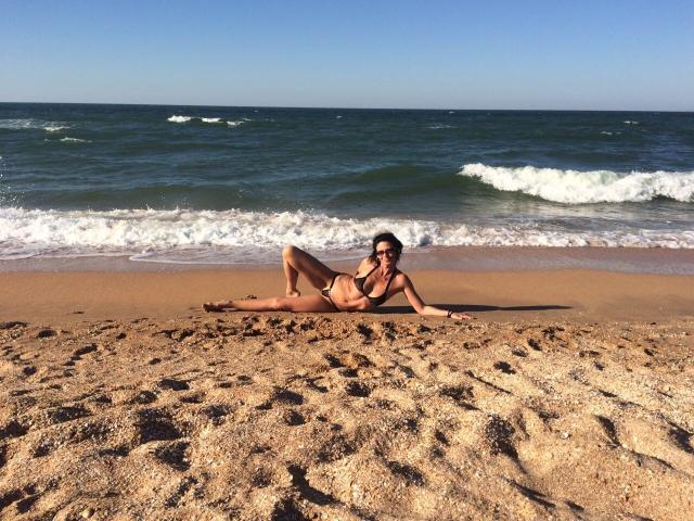 GannaWind, in costume da bagno, è sdraiata su una spiaggia sabbiosa con piccole onde, con i glutei rivolti verso l'alto, le gambe distese e le braccia appoggiate sulla sabbia. Sullo sfondo si vedono l'oceano blu e un cielo sereno..