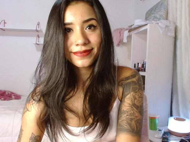 AshleyLover with long black hair, red lipstick, white tank top, tattooed arm, smiling at camera, pink bed in background, white shelves with makeup, roll of toilet paper.