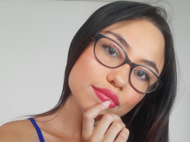 LolyFunX has black hair, glasses, pink lipstick, and is biting her finger with a cute smile, wearing a blue top, showing her soft, fair-skinned face.