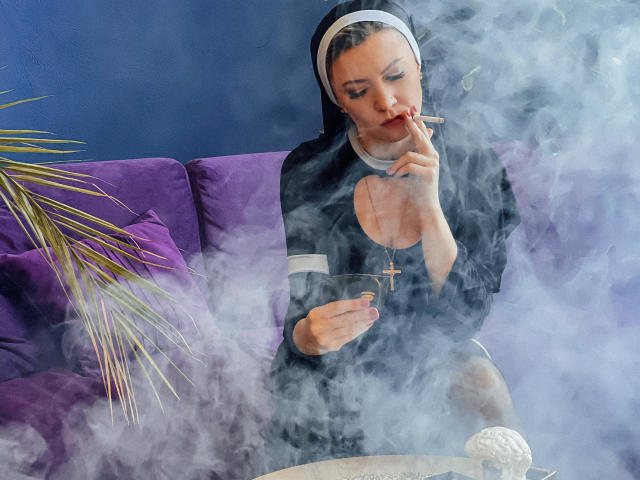 ParadiseRoxanne, eine Nonne mit einem schwarzen Habit und einer Kreuzkette, raucht einen Joint, umgeben von weißem Rauch, sitzend auf einem violetten Sofa und haltend eine Glaspfeife..