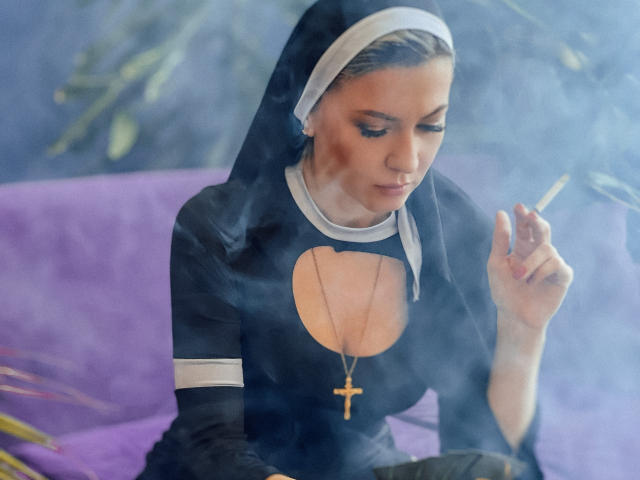 ParadiseRoxanne, uma freira com um hábito preto e um capuz branco, fuma um cigarro, segurando-o com a mão direita, usando um colar com uma cruz dourada, com fumaça azul ao redor..