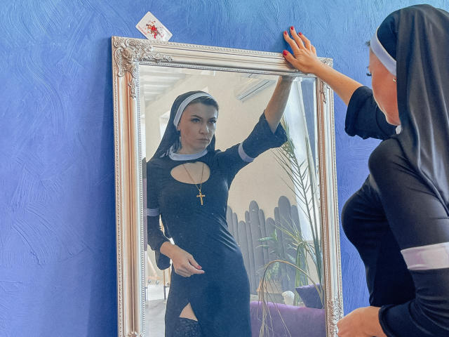 ParadiseRoxanne in un abito nero da suora con un taglio che mostra i suoi seni, indossa una collana con una croce, è in piedi di fronte a uno specchio e sta toccando la cornice. Parete blu..