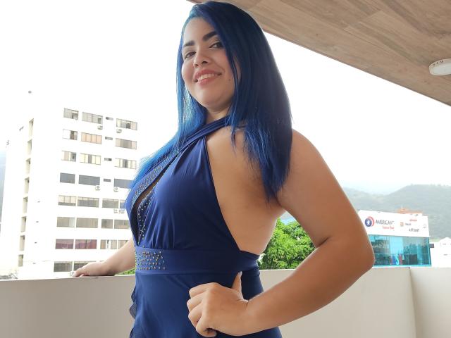 KimLafayette, avec ses cheveux bleus, portant une robe bleu foncé avec des découpes sur les côtés, souriante, montrant ses seins, debout avec une main sur sa hanche, sur un balcon..