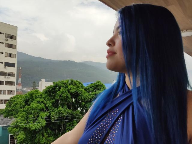 KimLafayette with blue hair, wearing a blue dress with gold studs, looking to the side, showing her nose and lips, standing near a roof, mountains in the background.