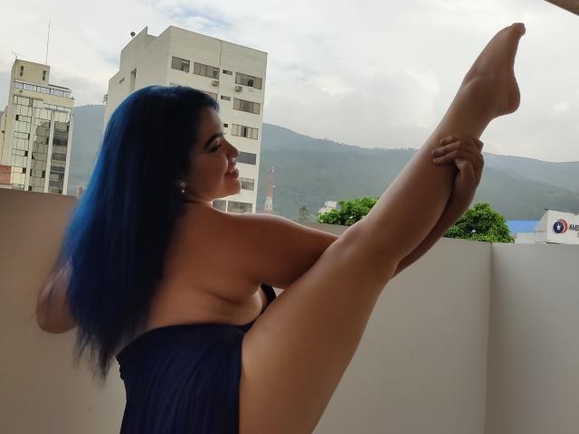 KimLafayette, avec ses cheveux bleus, nue à l'exception d'un tissu bleu foncé, levant une jambe, la tenant avec ses mains, montrant ses fesses et une vue latérale de ses seins..