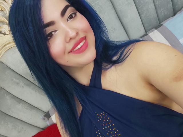 KimLafayette with long blue hair, smiling, wearing a dark blue halter top, showing bare shoulder, pink lipstick, and brown eyes, on a bed with gray and white pillows.